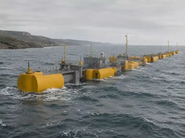 Eine lange Reihe großer gelber und grauer Wellenkraftwerk-Schwimmkörper liegt in kabbeligen Gewässern in der Nähe einer felsigen Küste unter einem bewölkten Himmel vor Anker.