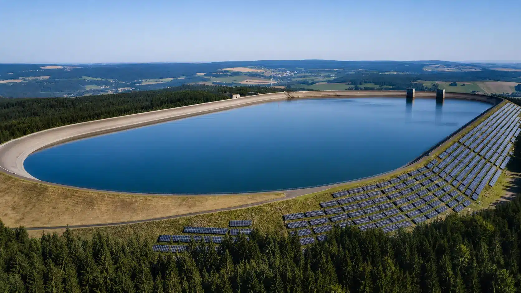 Ein großer, ovaler, von Bäumen umgebener künstlicher Stausee mit Reihen von Solarzellen, die auf einem grasbewachsenen Hang in der Nähe des Wassers installiert sind, stellt ein Speicherkraftwerk unter blauem Himmel in einer ruhigen ländlichen Landschaft dar.