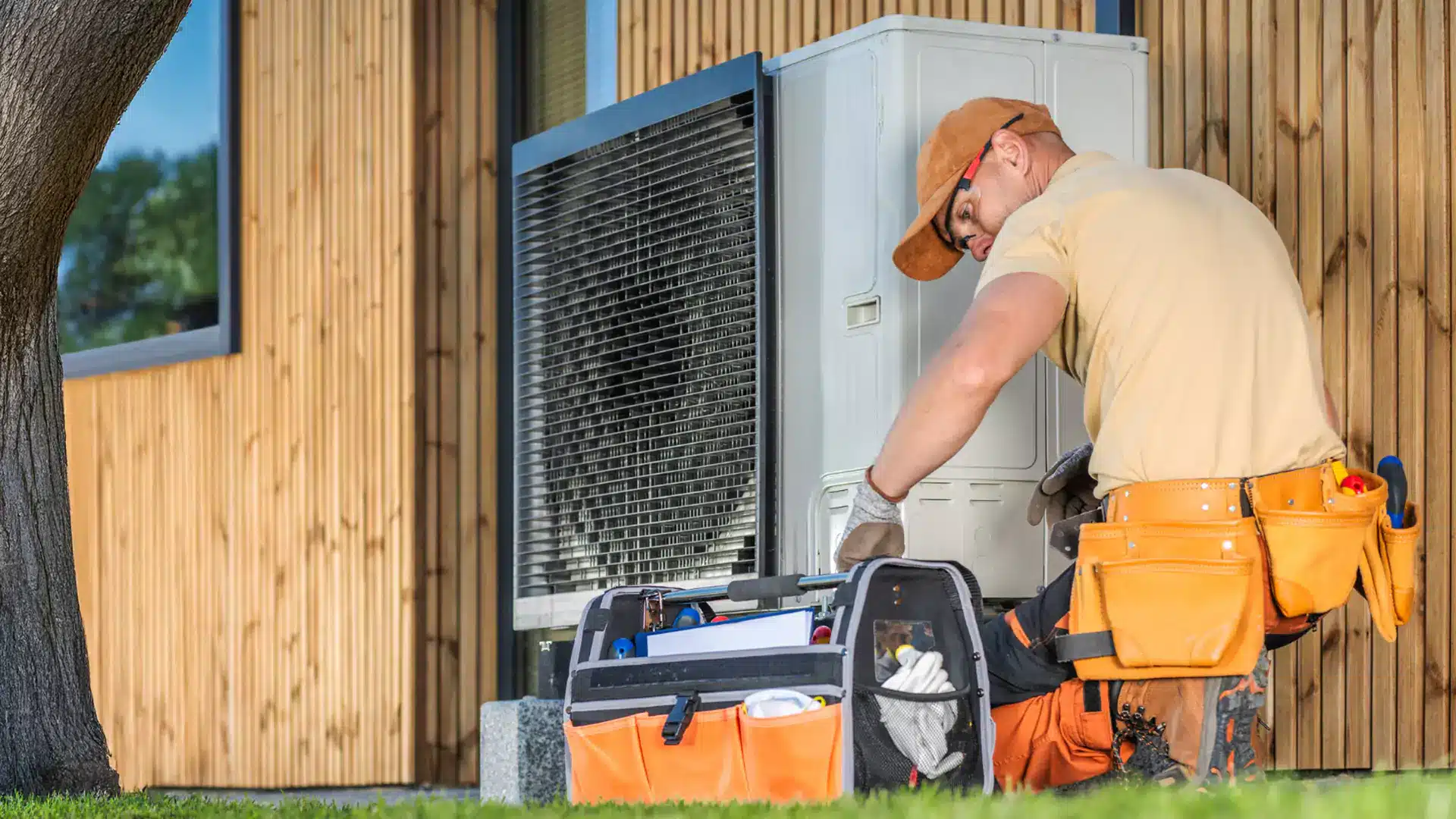 Ein Techniker mit orangefarbener Mütze und Werkzeuggürtel hockt neben einer Klimaanlage vor einem Holzgebäude. Er greift in eine schwarz-orangefarbene Werkzeugtasche und arbeitet an der Anlage. Möglicherweise nimmt er an einer Förderung für die energetische Sanierung teil, um die Energieeffizienz zu verbessern.