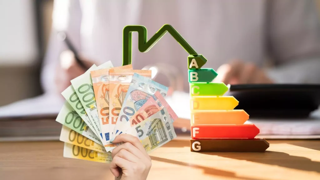 Im Vordergrund ist eine Hand zu sehen, die einen Fächer aus Euro-Banknoten hält. Dahinter ist ein 3D-Modell eines Energieeffizienzdiagramms in Form eines Hauses zu sehen, das bunte Balken mit den Beschriftungen A bis G zeigt. Im unscharfen Hintergrund arbeitet eine Person an einem Schreibtisch.