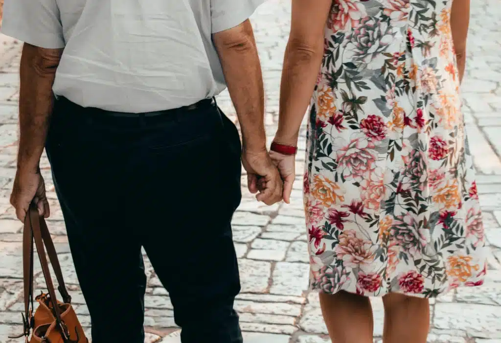 Ein Paar, von hinten gesehen, schlendert Hand in Hand über einen Kopfsteinpflasterweg. Der Mann, konzentriert auf den Vermögensaufbau mit 50, trägt ein weißes Hemd, eine dunkle Hose und eine Tasche. Die Frau wirkt lebhaft und trägt ein farbenfrohes Blumenkleid. Ihre Hände greifen sanft in die Mitte ihres Weges.