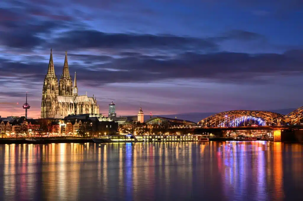 Eine nächtliche Stadtansicht von Köln, Deutschland, mit dem beleuchteten Kölner Dom und der Hohenzollernbrücke über dem Rhein – eine inspirierende Szene für alle, die inmitten der farbenfrohen Spiegelungen und des dramatischen Himmels der Stadt nach einem Stromvergleich Köln suchen.
