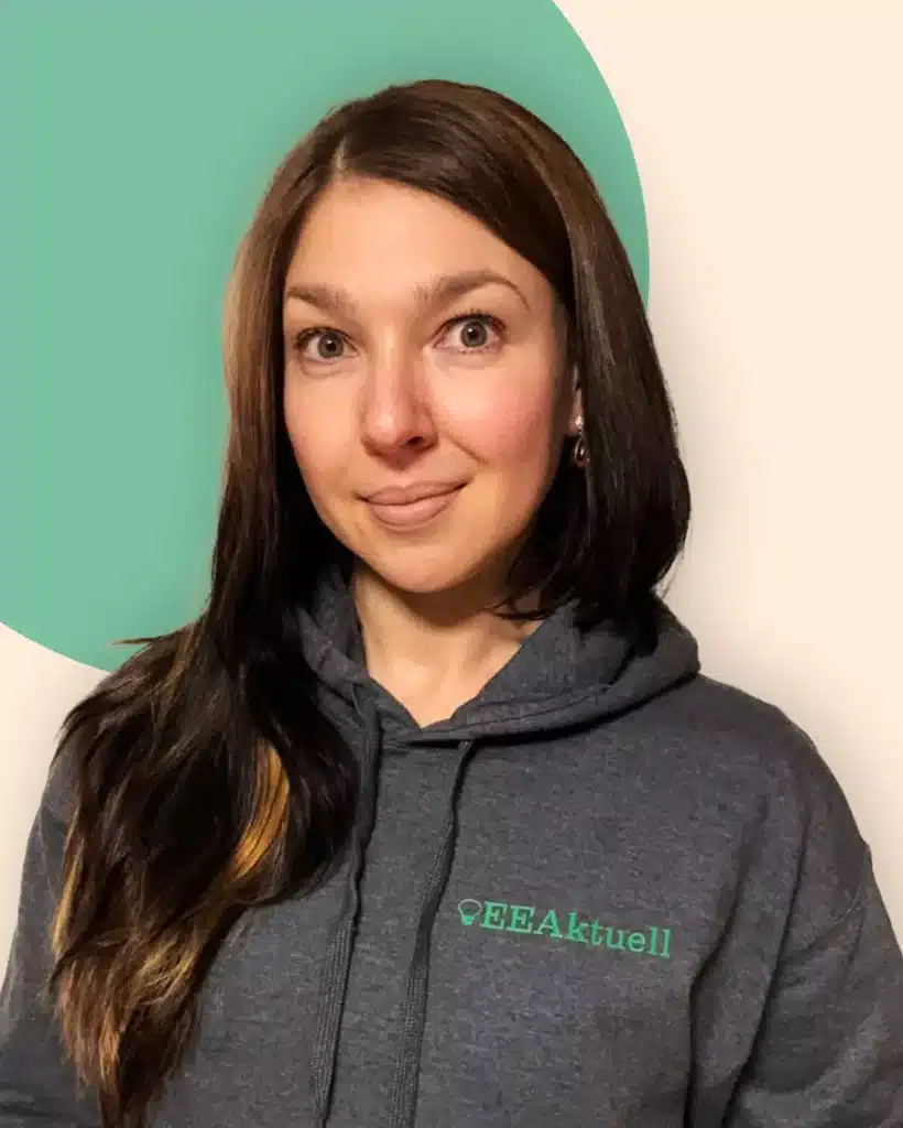 Eine Frau mit langen braunen Haaren, die einen grauen Kapuzenpulli mit der Aufschrift EEAktuell und Erneuerbare Energien trägt, steht vor einem beigen Hintergrund mit einem grünen Kreis hinter ihr. Sie schaut in die Kamera und lächelt leicht.