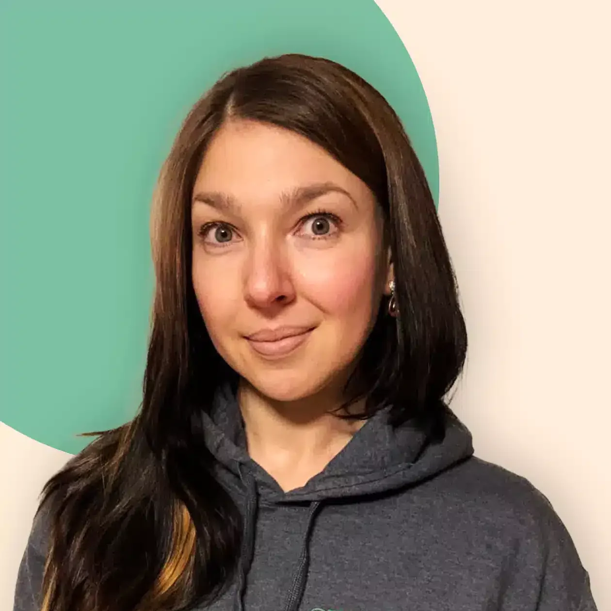 Eine Frau mit langen braunen Haaren, die einen grauen Kapuzenpulli mit der Aufschrift EEAktuell und Erneuerbare Energien trägt, steht vor einem beigen Hintergrund mit einem grünen Kreis hinter ihr. Sie schaut in die Kamera und lächelt leicht.