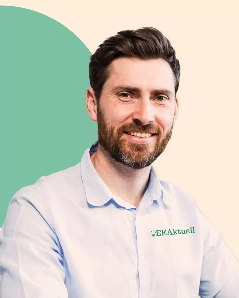 Ein Mann mit braunen Haaren und Bart, der ein hellblaues QEEAktuell-Shirt trägt, lächelt vor einem cremefarbenen und grünen Hintergrund, der seine Begeisterung für Erneuerbare Energien widerspiegelt.
