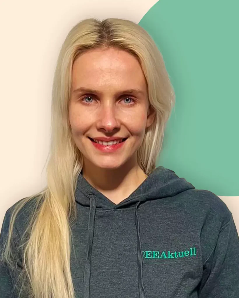 Eine Frau mit langen blonden Haaren lächelt in die Kamera. Sie trägt einen grauen Kapuzenpullover mit dem Aufdruck PEEAktuell, der ihr Interesse an Erneuerbaren Energien widerspiegelt. Der Hintergrund ist hell mit einem grünen, runden Akzent.