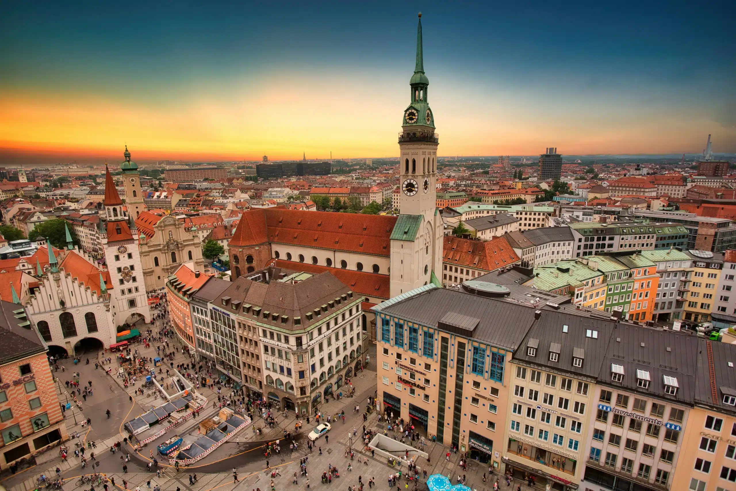 Luftaufnahme des Münchner Marienplatzes bei Sonnenuntergang mit historischen Gebäuden, dem Turm der Peterskirche, farbenfrohen Dächern und Menschenmassen, die sich auf dem belebten Stadtplatz versammeln - ein Energiezentrum, das sich perfekt für einen stromvergleich München eignet.
