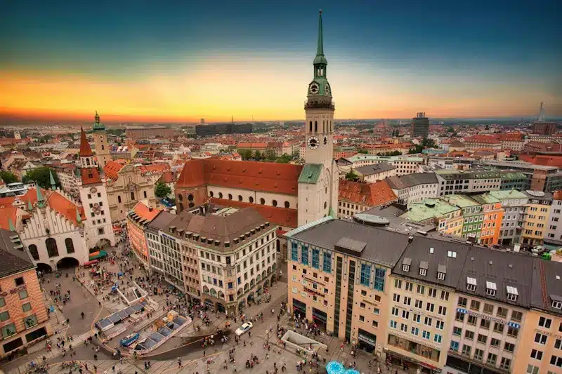 Luftaufnahme des Münchner Marienplatzes bei Sonnenuntergang mit historischen Gebäuden, dem Turm der Peterskirche, farbenfrohen Dächern und Menschenmassen, die sich auf dem belebten Stadtplatz versammeln - ein Energiezentrum, das sich perfekt für einen stromvergleich München eignet.