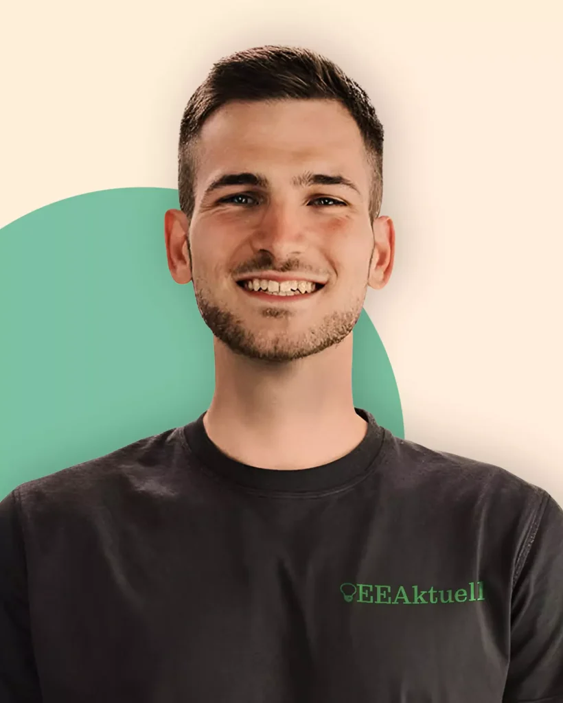 Ein junger Mann mit kurzen braunen Haaren und heller Gesichtsbehaarung lächelt in die Kamera. Er trägt ein dunkles T-Shirt mit der Aufschrift "EEAktuell" - eine Anspielung auf Erneuerbare Energien. Der Hintergrund ist hell mit einem grünen Halbkreis hinter ihm.