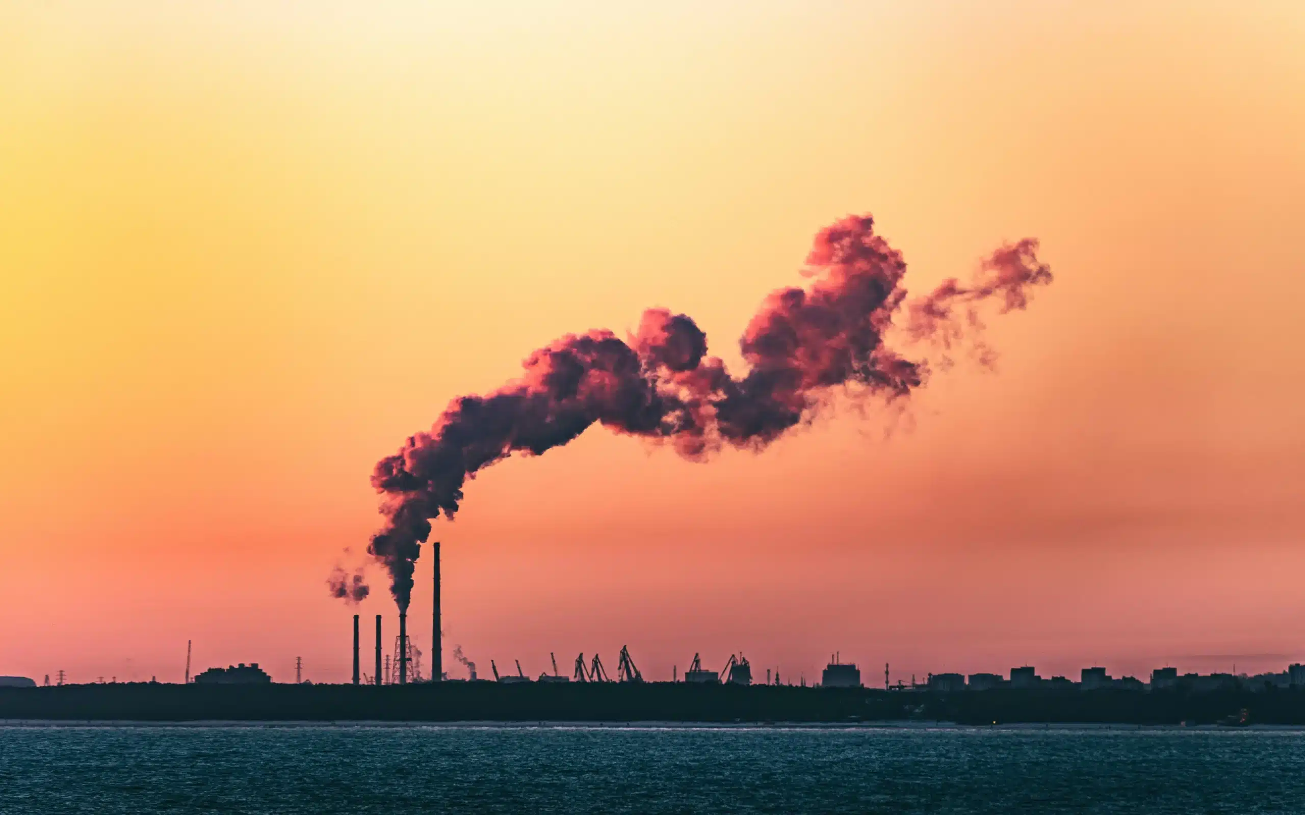 Fabrikschornsteine, die dichten, dunklen Rauch vor einem orangefarbenen und rosafarbenen Himmel bei Sonnenuntergang ausstoßen, mit Wasser im Vordergrund und Industriegebäuden als Silhouetten im Hintergrund - eine überzeugende Szene, die zum Verzicht auf fossile Brennstoffe auffordert.