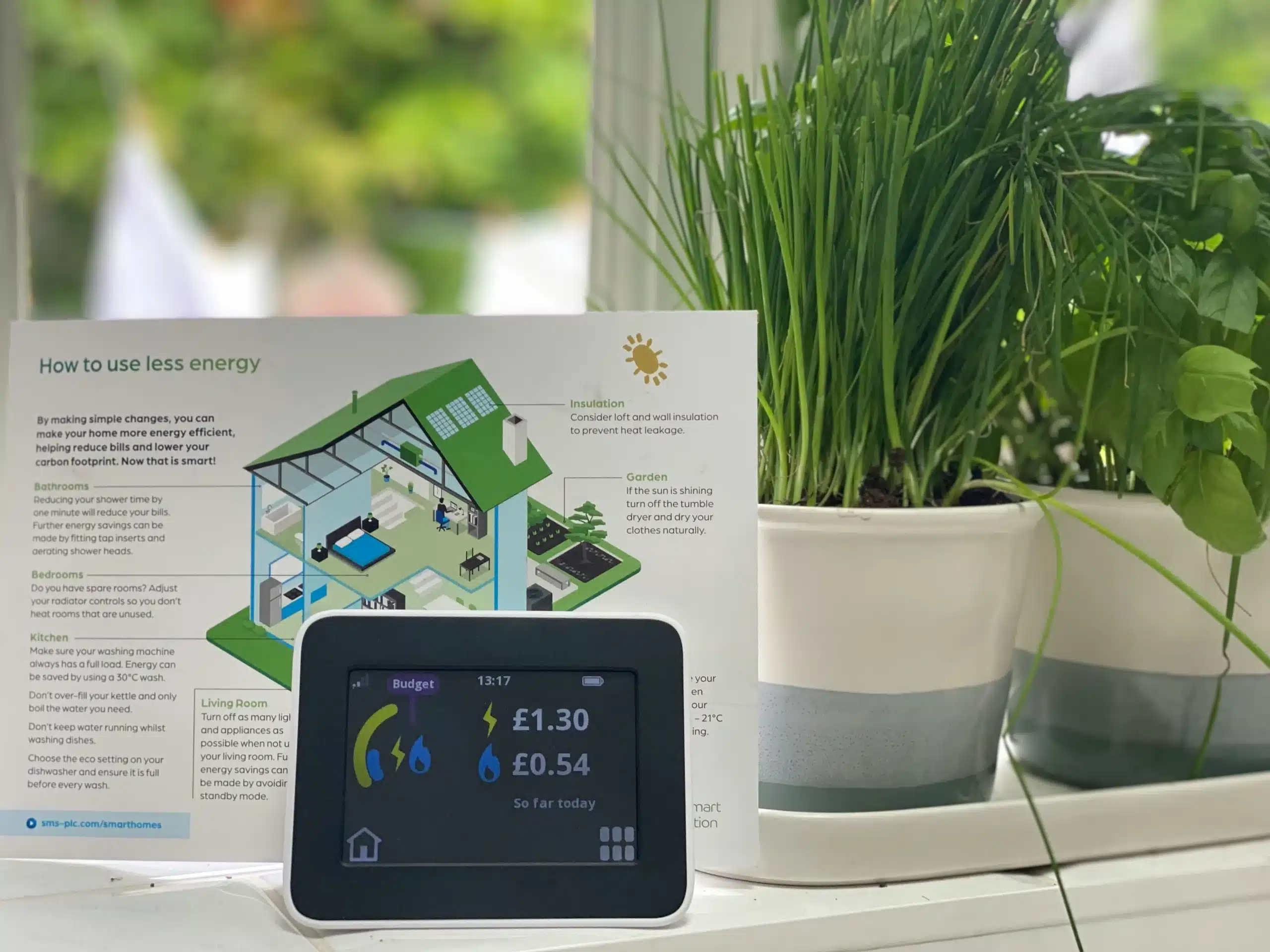Ein intelligenter Stromzähler zeigt Verbrauch und Kosten auf einer Fensterbank neben grünen Kräutertöpfen an. Dahinter befindet sich ein illustriertes Faltblatt zur Senkung des Energieverbrauchs mit hilfreichen Tipps und einem Diagramm für den Haushalt.