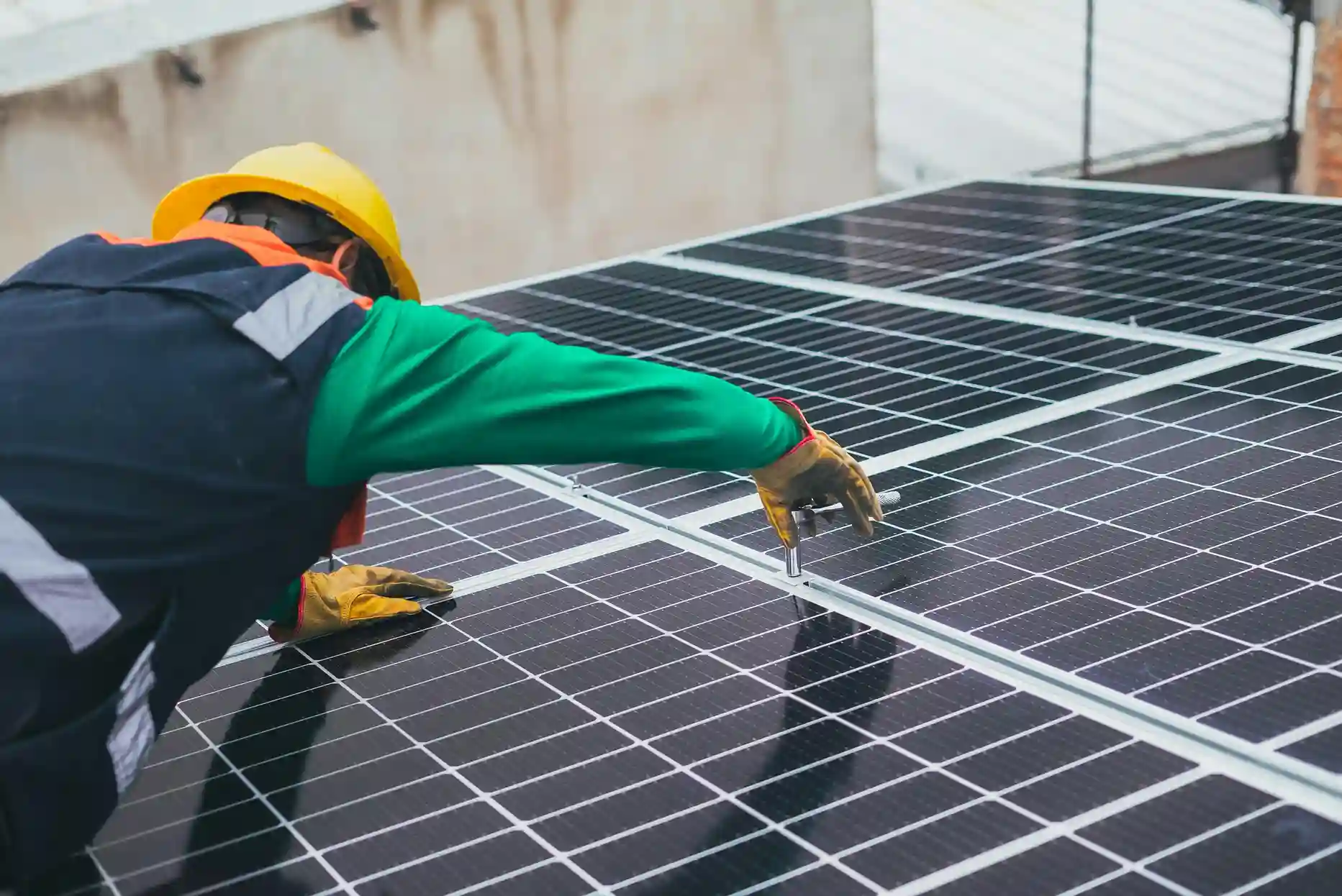 Ein Arbeiter, der einen gelben Schutzhelm und eine Sicherheitsweste trägt, installiert oder wartet Solarmodule auf einem Dach und benutzt ein Werkzeug, um die Befestigungen der Module zu justieren - ein Beispiel dafür, wie eine Energiegenossenschaft nachhaltige Energielösungen unterstützen kann.