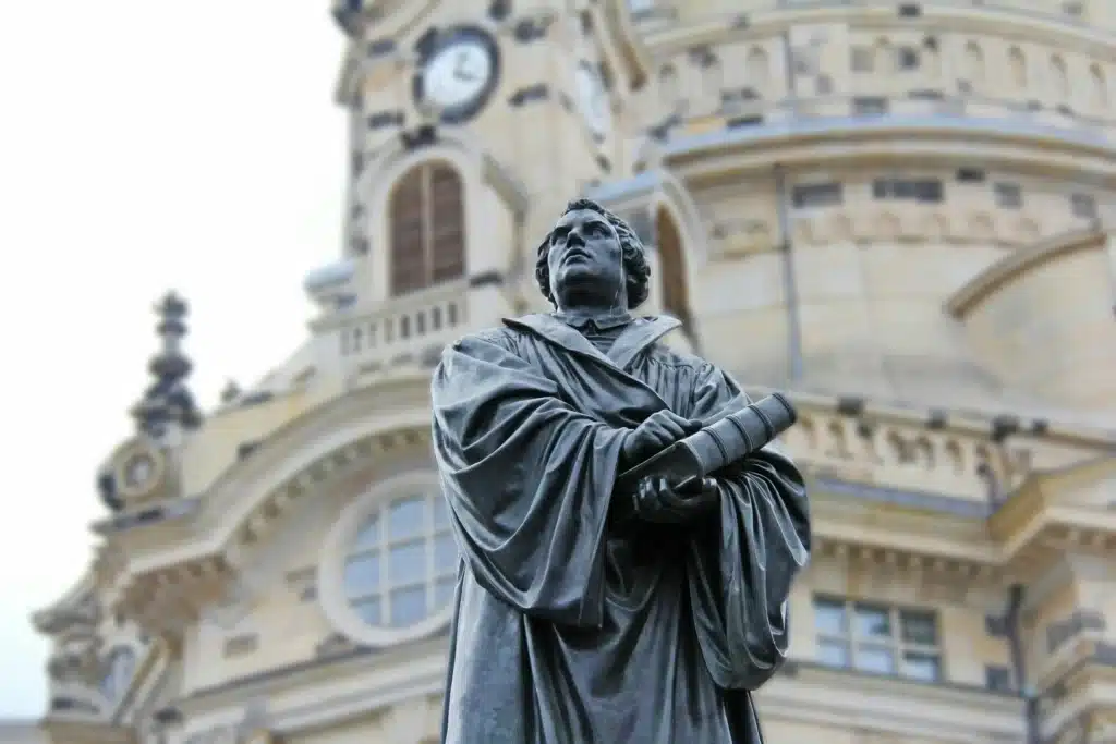 Die Bronzestatue eines Mannes in akademischer Robe, der eine Schriftrolle in der Hand hält, steht vor einem kunstvollen, historischen Gebäude mit Uhr und Bogenfenstern - ein ikonischer Anblick in Dresden, nur wenige Schritte von dem Ort entfernt, an dem Einheimische nach dem besten Stromvergleich suchen, den Dresden zu bieten hat.