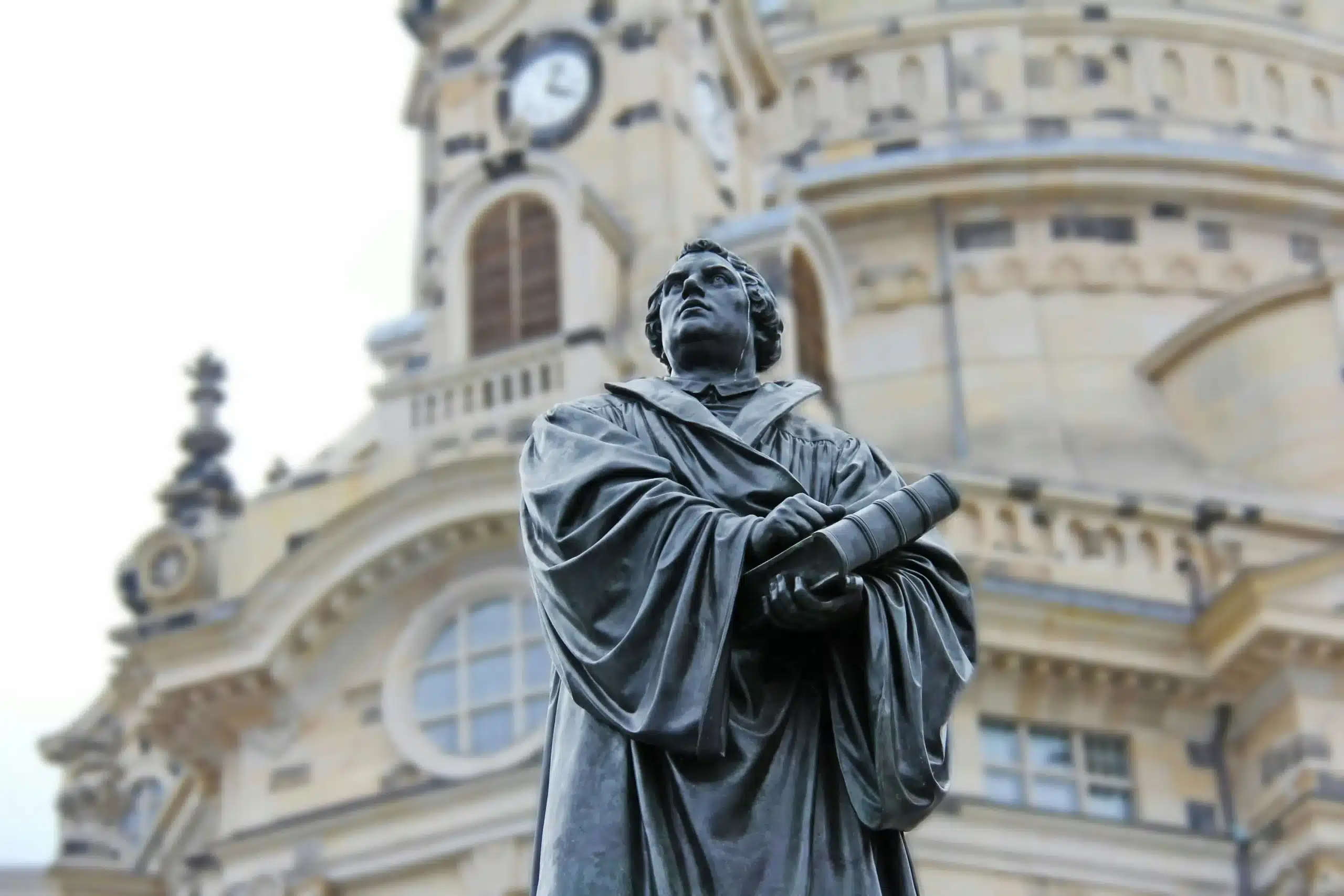 Die Bronzestatue eines Mannes in akademischer Robe, der eine Schriftrolle in der Hand hält, steht vor einem kunstvollen, historischen Gebäude mit Uhr und Bogenfenstern - ein ikonischer Anblick in Dresden, nur wenige Schritte von dem Ort entfernt, an dem Einheimische nach dem besten Stromvergleich suchen, den Dresden zu bieten hat.