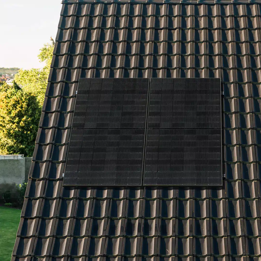 Zwei schwarze Sonnenkollektoren sind nebeneinander auf einem dunklen, mit Ziegeln gedeckten Schrägdach montiert. Im Hintergrund sind an einem sonnigen Tag Bäume, Gras und ein Teil einer Mauer zu sehen.