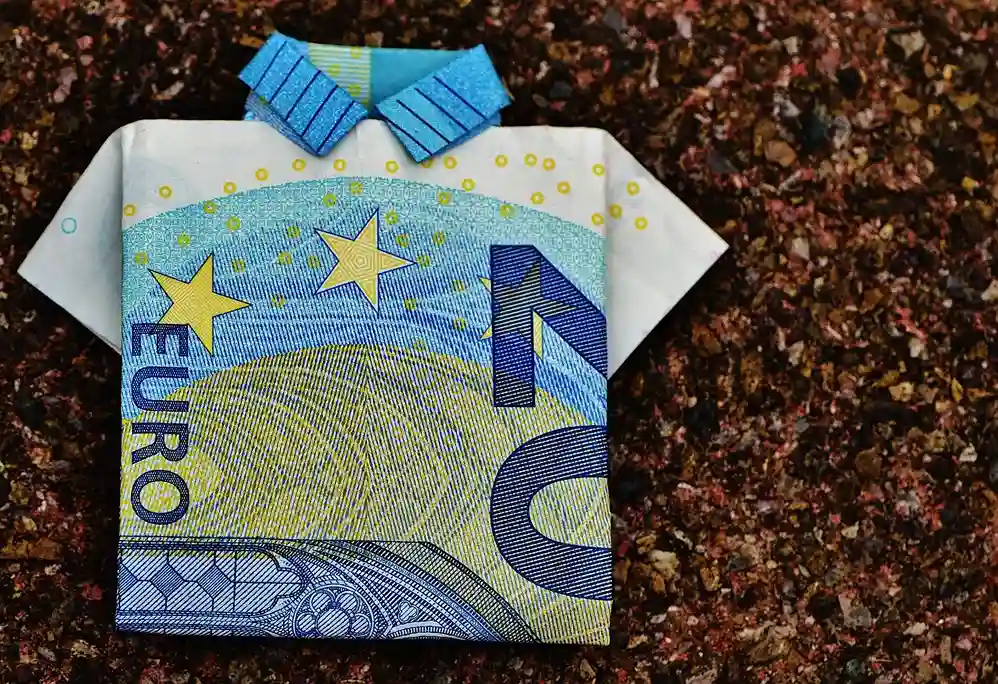 Ein Zehn-Euro-Schein, gefaltet in die Form eines Hemdes mit Kragen, platziert auf einer strukturierten braunen Oberfläche - die perfekte Inspiration, wenn Sie überlegen, vielleicht ein Tagesgeldkonto zu eröffnen.