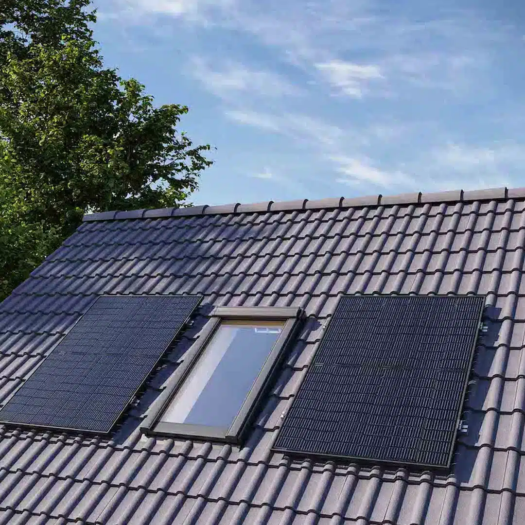 Zwei Solarmodule sind auf einem mit Ziegeln gedeckten Schrägdach neben einem Dachfenster installiert, mit einem Baum und einem klaren blauen Himmel im Hintergrund.