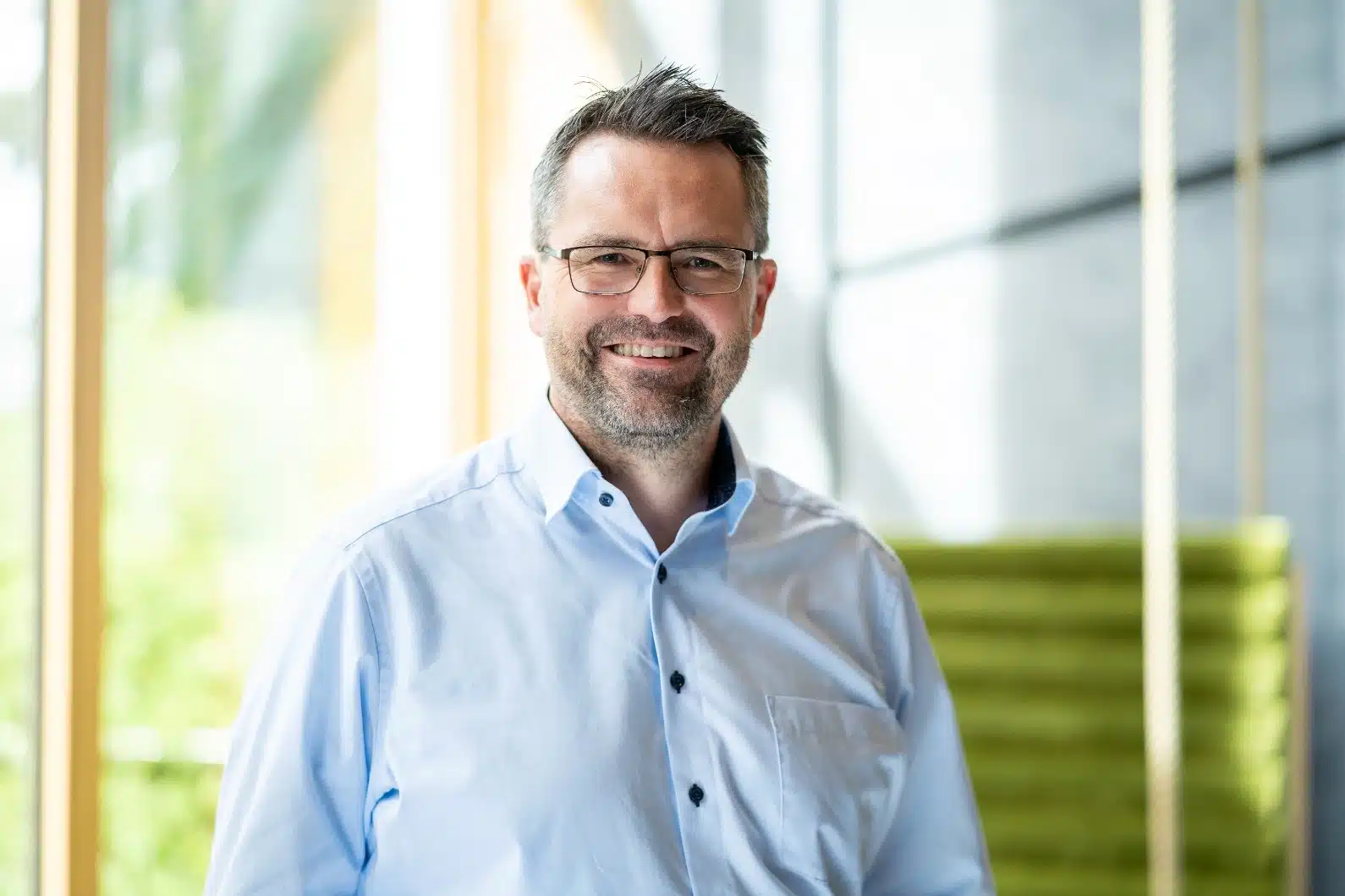 Ein Mann mit kurzen braunen Haaren, Brille und Bart lächelt, während er ein hellblaues Button-up-Hemd trägt. Er steht in einem Innenraum des Büros von GLS Investments mit großen Fenstern und grünen Sitzgelegenheiten im Hintergrund.