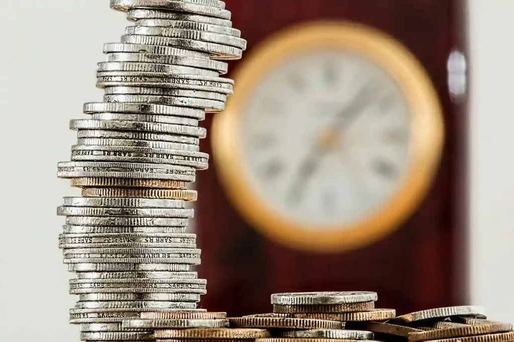 Im Vordergrund ist ein hoher, ungleichmäßiger Stapel Silbermünzen zu sehen, im Hintergrund eine unscharfe Uhr, die das Konzept von Zeit und Geld veranschaulicht - ideal für die Diskussion von Tagesgeld- und Festgeldoptionen.
