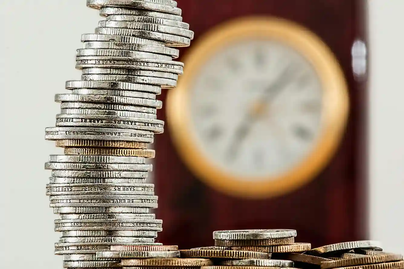 Im Vordergrund ist ein hoher, ungleichmäßiger Stapel Silbermünzen zu sehen, im Hintergrund eine unscharfe Uhr, die das Konzept von Zeit und Geld veranschaulicht - ideal für die Diskussion von Tagesgeld- und Festgeldoptionen.
