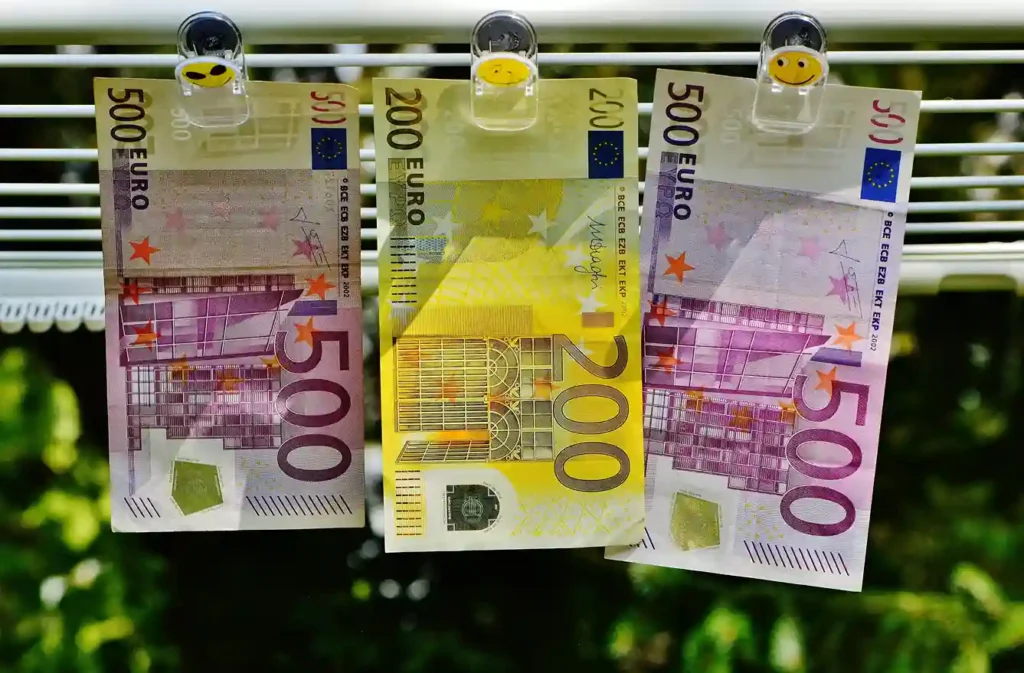 Drei Euro-Banknoten (zwei 500er und ein 200er) hängen auf einer Linie mit Smiley-Klammern, die an Geldwäsche erinnern. Der unscharfe grüne Hintergrund deutet die Natur an - ein Bild, das so kontrastreich ist wie die Wahl zwischen Tagesgeld und Festgeld für Ihre Ersparnisse.