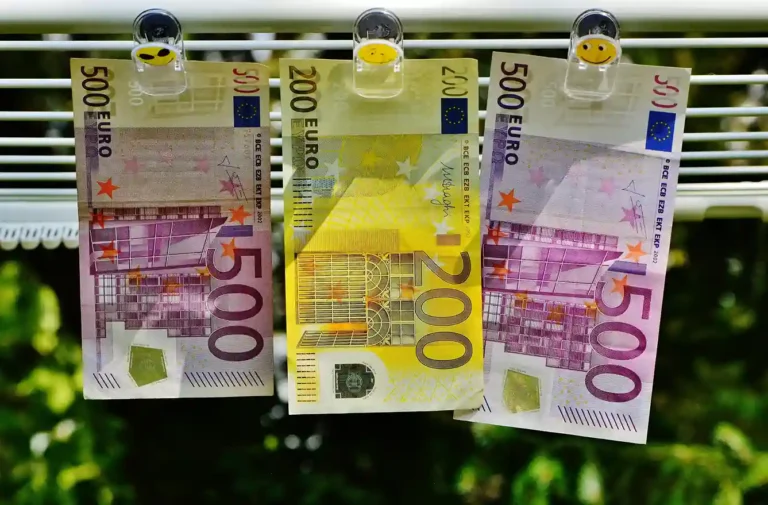 Drei Euro-Banknoten (zwei 500er und ein 200er) hängen auf einer Linie mit Smiley-Klammern, die an Geldwäsche erinnern. Der unscharfe grüne Hintergrund deutet die Natur an - ein Bild, das so kontrastreich ist wie die Wahl zwischen Tagesgeld und Festgeld für Ihre Ersparnisse.