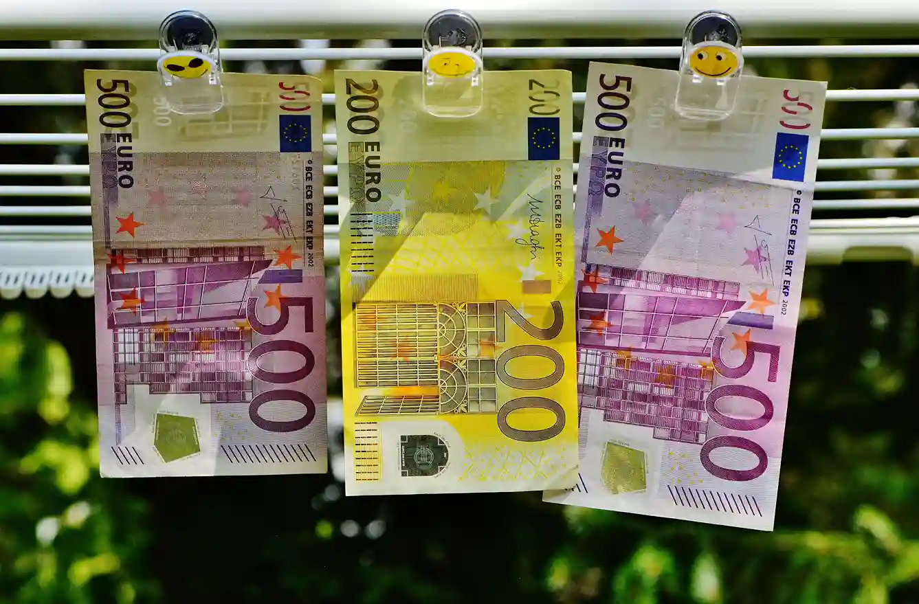 Drei Euro-Banknoten (zwei 500er und ein 200er) hängen auf einer Linie mit Smiley-Klammern, die an Geldwäsche erinnern. Der unscharfe grüne Hintergrund deutet die Natur an - ein Bild, das so kontrastreich ist wie die Wahl zwischen Tagesgeld und Festgeld für Ihre Ersparnisse.