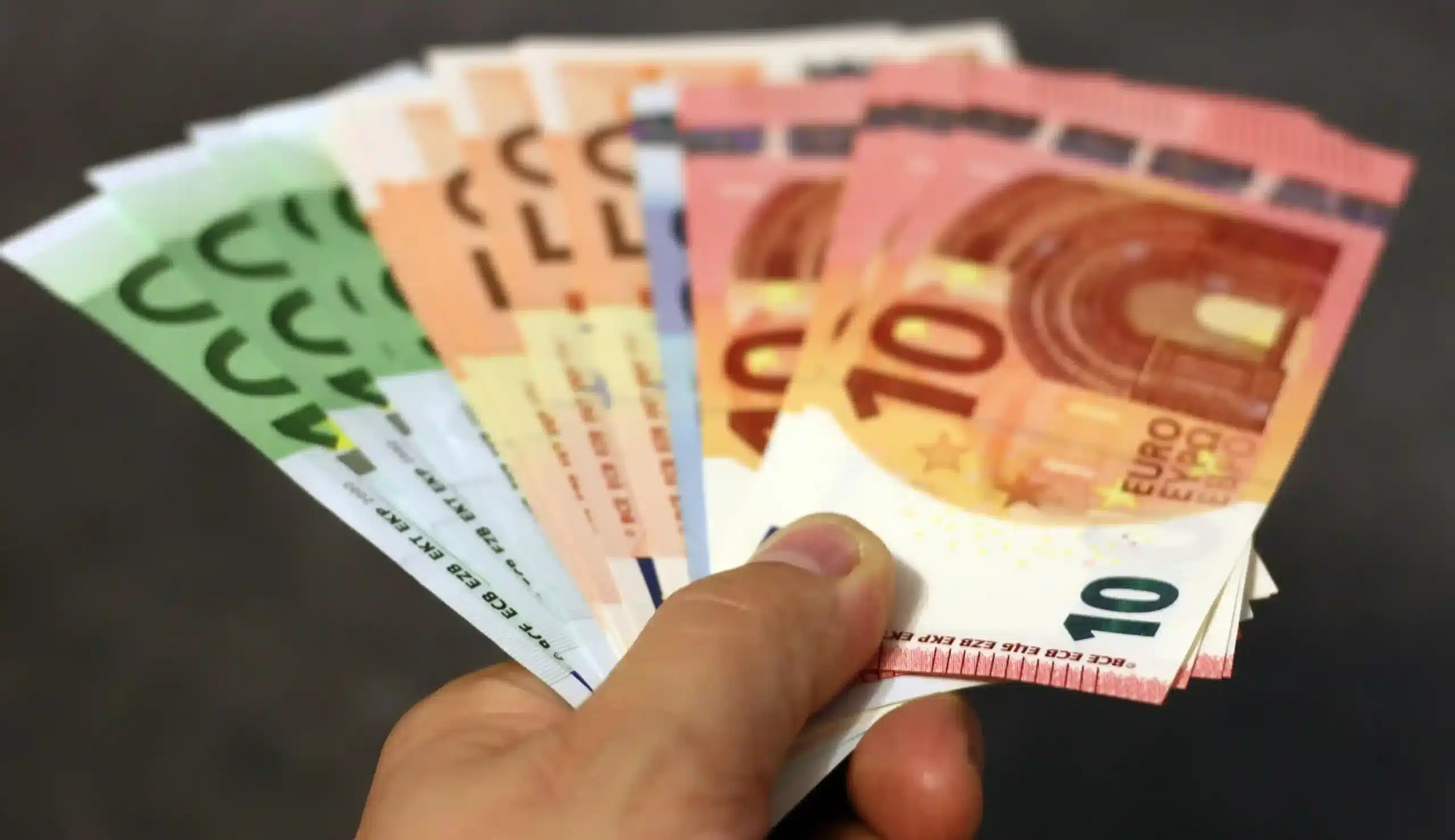 Eine Hand, die einen Fächer von Euro-Banknoten in verschiedenen Stückelungen hält, darunter 5, 10, 20 und 50 Euro - ein Bild, das für Ersparnisse oder eine alternative Festgeldoption stehen könnte - vor einem verschwommenen Hintergrund.