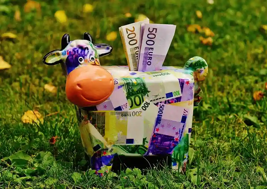 Ein farbenfrohes Sparschwein in Form einer Kuh, die mit Euro-Banknoten bedruckt ist, steht auf einer Wiese. Aus seinem Rücken ragen 500- und 200-Euro-Scheine heraus und symbolisieren die Wahl zwischen Tagesgeld und Festgeld beim Sparen.