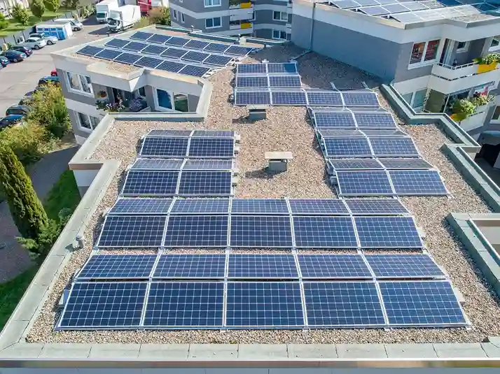 Luftaufnahme eines flachen, mit mehreren Solarmodulen bedeckten Daches eines modernen Gebäudes, das die Vorteile von Energiegenossenschaften gegenüber nachhaltigen Geldanlagen hervorhebt, umgeben von anderen Gebäuden, parkenden Autos und sichtbaren Grünflächen.
