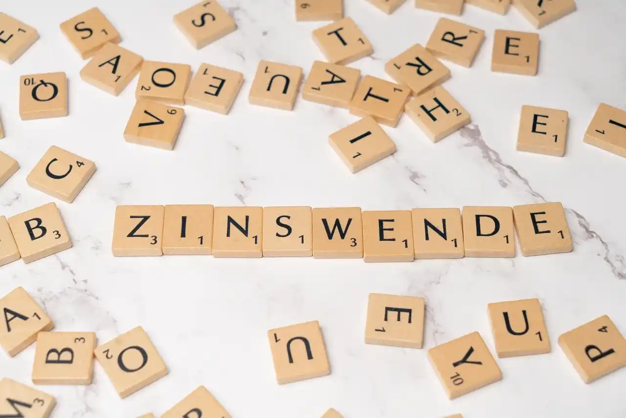 Scrabble-Buchstabenfliesen, die so angeordnet sind, dass sie in der Mitte "ZINSWENDE" buchstabieren, umgeben von verstreuten, zufälligen Fliesen auf einer weißen Marmoroberfläche - eine künstlerische Anspielung auf Finanzthemen wie Tagesgeld und Festgeld.