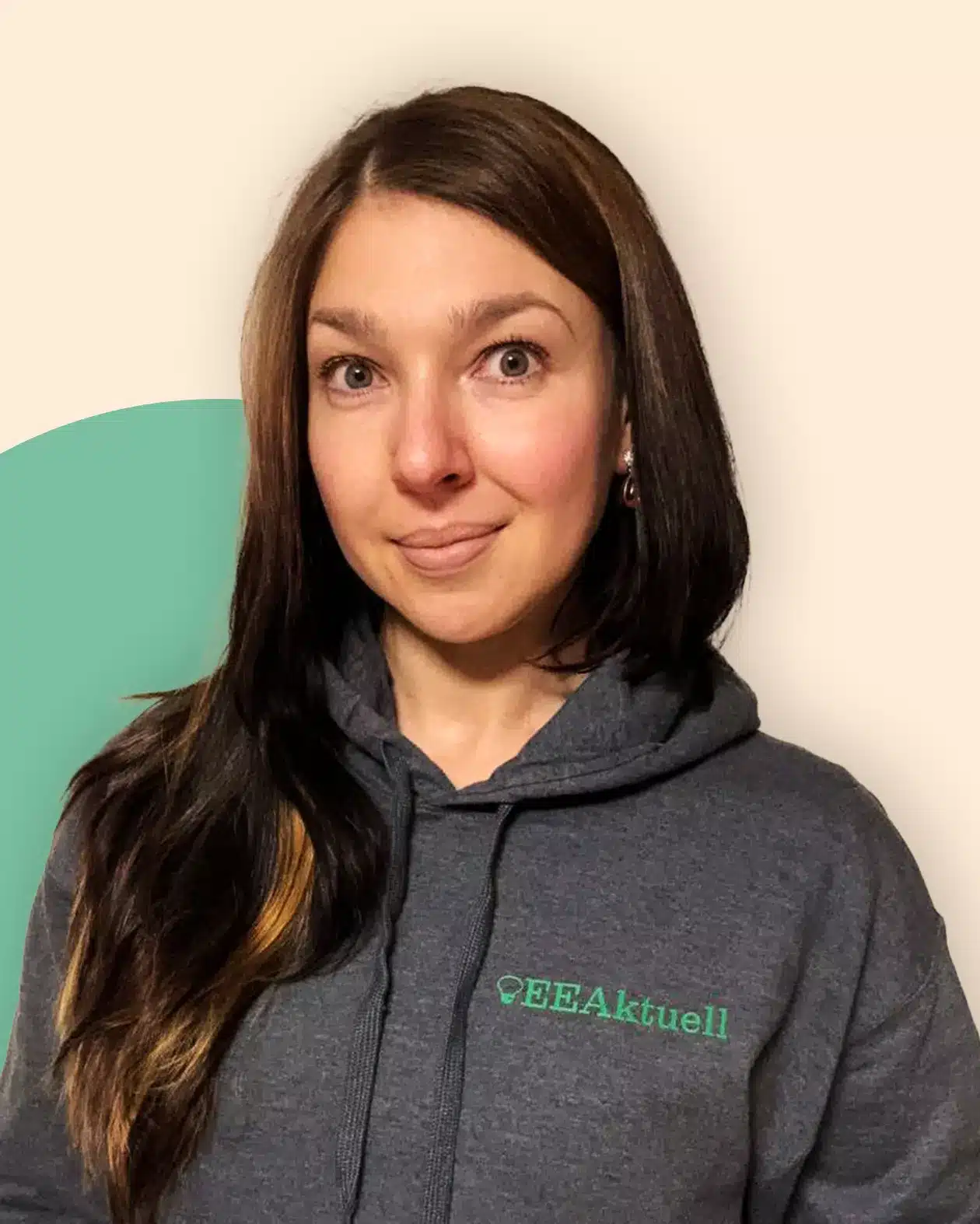 Eine Frau mit langen braunen Haaren, die einen grauen Kapuzenpullover mit der grünen Aufschrift EEAktuell trägt, steht vor einem cremefarbenen Hintergrund und einer grünen Form. Sie schaut in die Kamera und lächelt leicht, was ihr Interesse an Erneuerbaren Energien widerspiegelt.