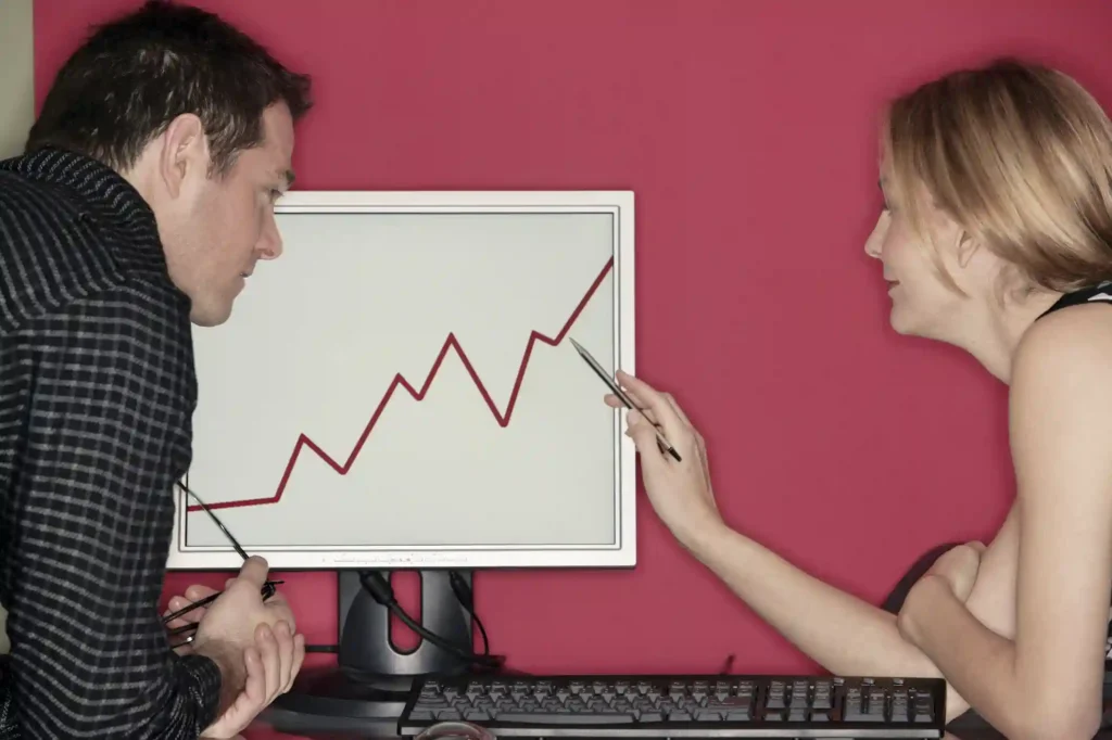 Ein Mann und eine Frau diskutieren über ein Liniendiagramm, das einen Aufwärtstrend auf einem Computermonitor zeigt - vielleicht um ihre justtrade erfahrung zu überprüfen. Die Frau zeigt mit einem Stift auf den Bildschirm, während der Mann zuhört und seine Brille festhält. Der Hintergrund ist einfarbig rosa-rot.