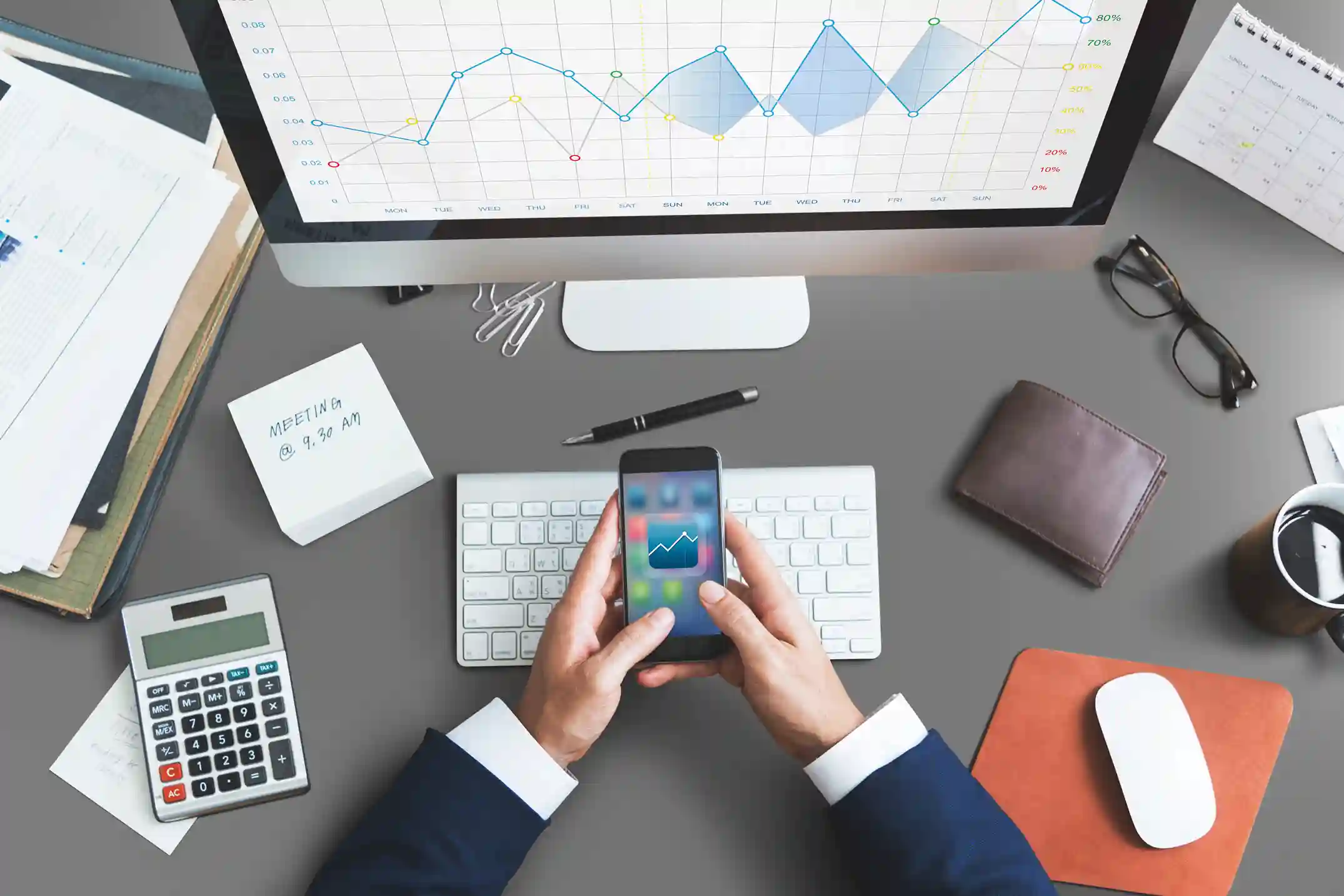 Eine Person in Geschäftskleidung, die ein Smartphone mit einem Diagramm in der Hand hält, sitzt an einem Schreibtisch mit einer Tastatur, einem Monitor mit einem Liniendiagramm, einem Taschenrechner, einem Notizblock, einer Brille, einer Brieftasche, einem Stift und einer Maus - perfekt, um eine positive justtrade erfahrung zu illustrieren.