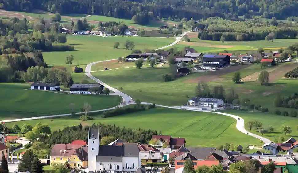 Eine kurvenreiche Landstraße schlängelt sich durch eine üppig grüne Landschaft mit verstreuten Häusern, Bauernhöfen, Feldern und Bäumen, alles vor dem Hintergrund bewaldeter Hügel - und spiegelt den Geist der Energiewende in dem friedlichen Dorf mit seiner malerischen Kirche im Vordergrund wider.