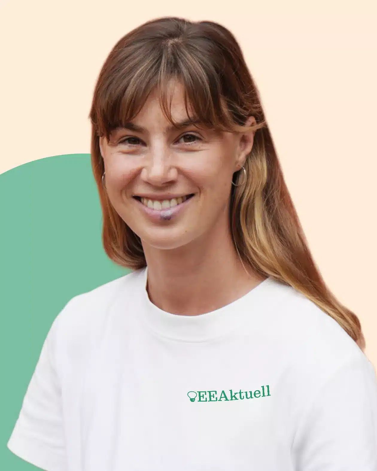Eine Frau mit hellbraunem Haar und Pony lächelt in die Kamera. Sie trägt ein weißes T-Shirt mit dem Aufdruck "EEAktuell", der ihr Interesse an Erneuerbaren Energien zum Ausdruck bringt. Der Hintergrund ist in hellen Pfirsich- und Grüntönen gehalten.