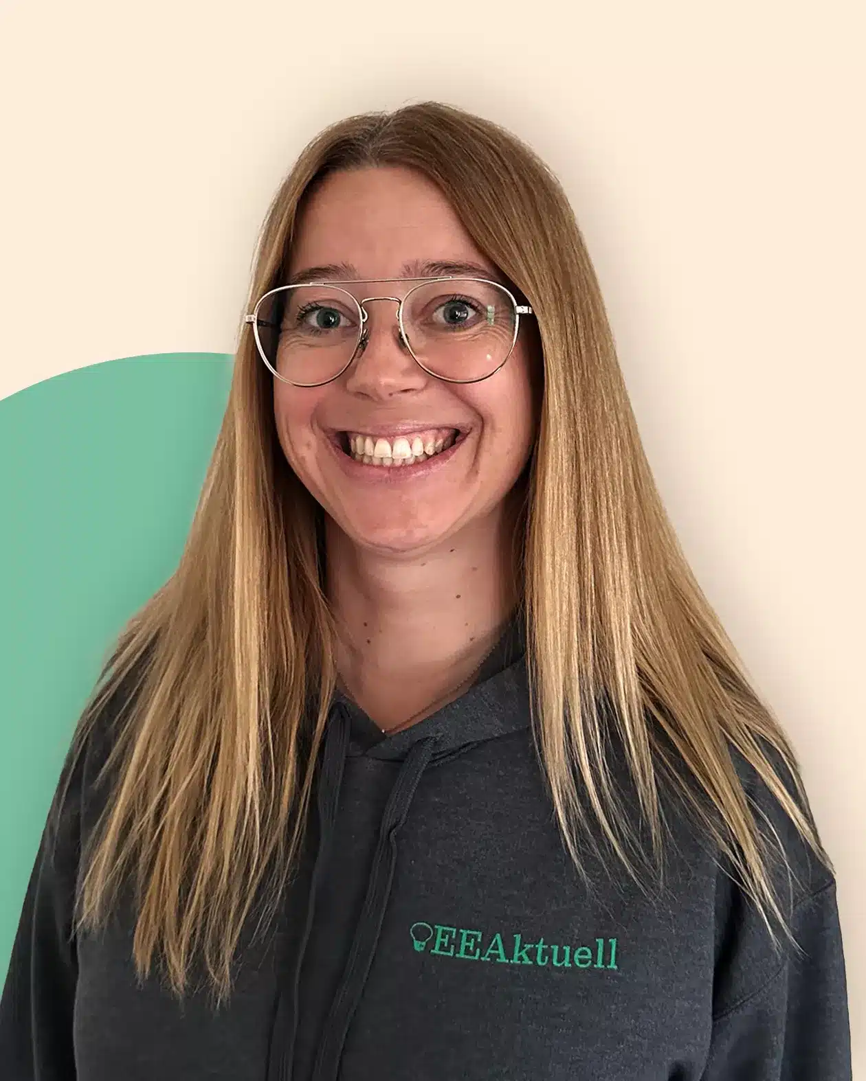 Eine lächelnde Frau mit langen blonden Haaren und Brille trägt einen grauen Kapuzenpullover mit dem Text EEAktuell, der Erneuerbare Energien hervorhebt. Der Hintergrund ist beige mit einer grünen geschwungenen Form auf der linken Seite.