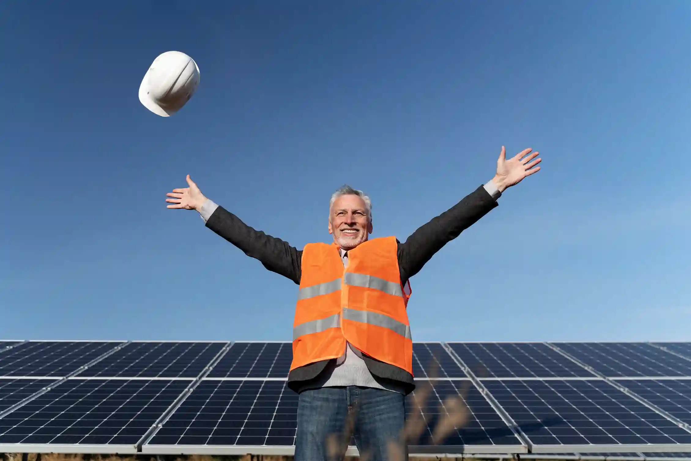 Ein lächelnder Mann in einer orangefarbenen Sicherheitsweste steht vor Sonnenkollektoren, hebt freudig die Arme und feiert positive Energiegenossenschaftserfahrungen, während er unter einem klaren blauen Himmel einen weißen Schutzhelm in die Luft wirft.