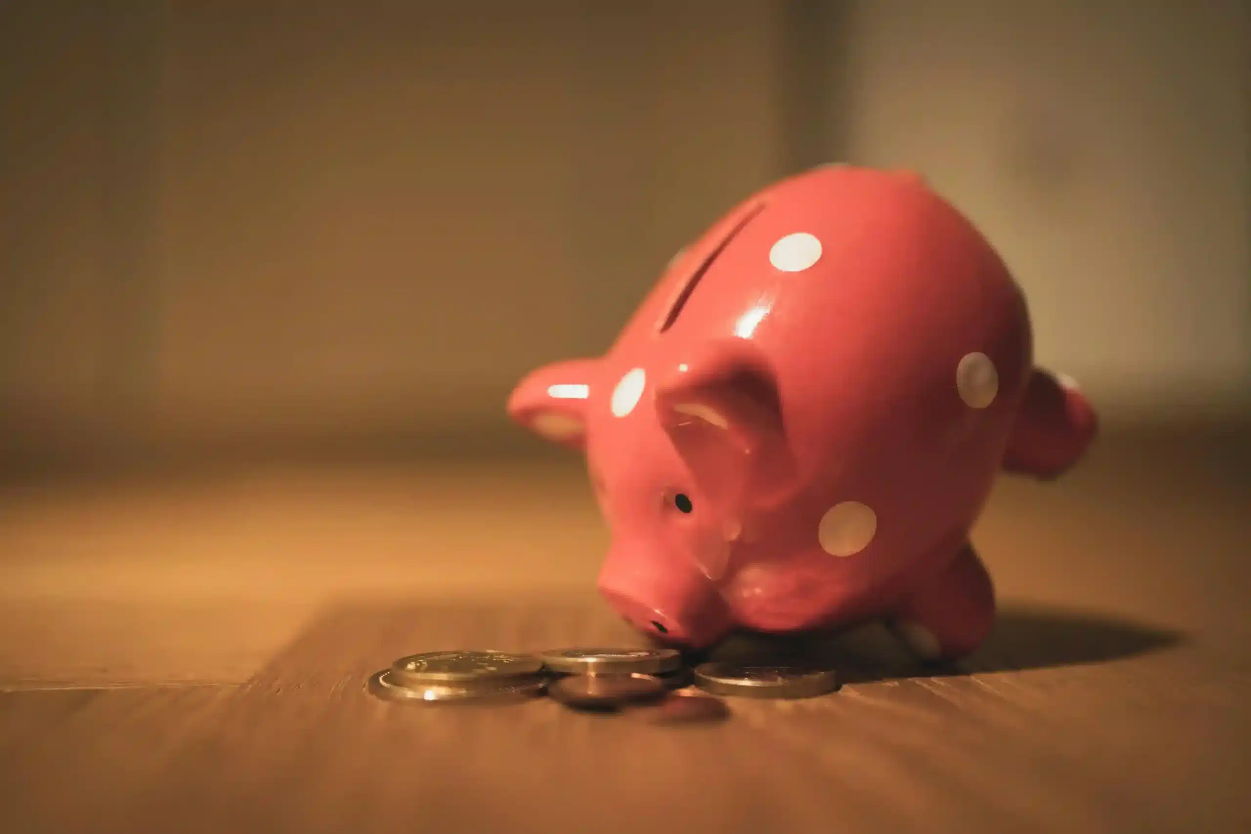 Ein rosafarbenes Sparschwein mit weißen Flecken ist nach vorne geneigt, als ob es einen kleinen Haufen Münzen beschnuppert oder vergleicht - ähnlich wie bei der Abwägung zwischen Tagesgeld und Festgeld. Die hölzerne Oberfläche und der sanft verschwommene Hintergrund geben einen gemütlichen Ton an.