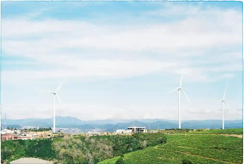 Drei weiße Windräder stehen auf einem grünen Hügel mit einigen Gebäuden und symbolisieren die Energiewende unter einem blauen Himmel mit leichten Wolken und fernen Bergen im Hintergrund.