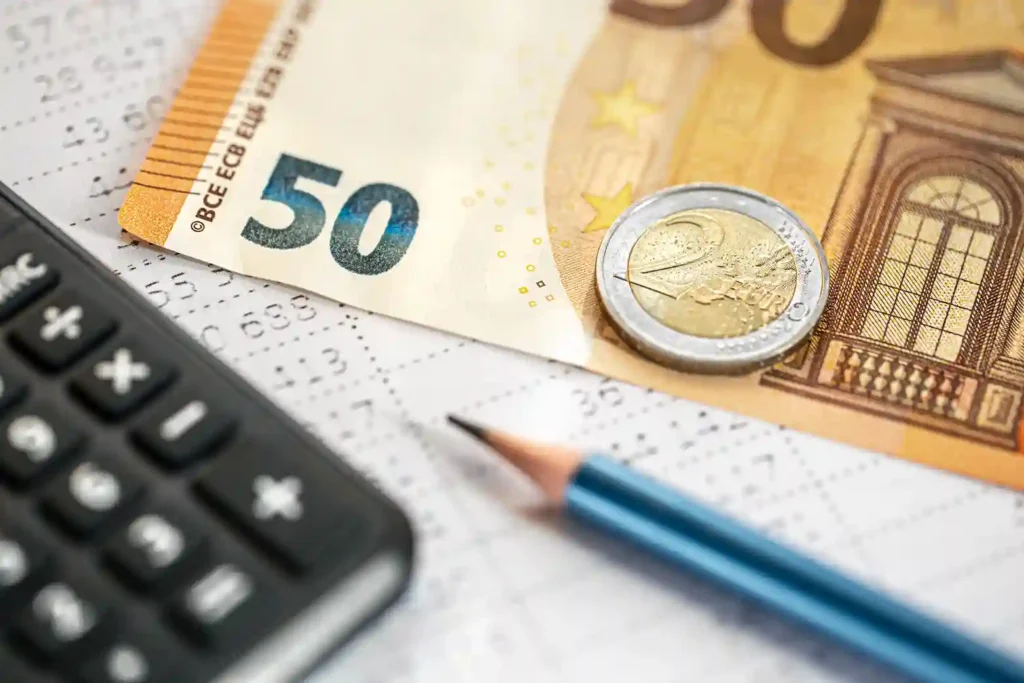 Eine Nahaufnahme einer 50-Euro-Banknote, einer 2-Euro-Münze, eines Bleistifts und eines Taschenrechners - ideale Werkzeuge für die Verwendung eines Zinsrechner-Festgelds -, die auf Finanzdokumenten mit aufgedruckten Zahlen liegen.