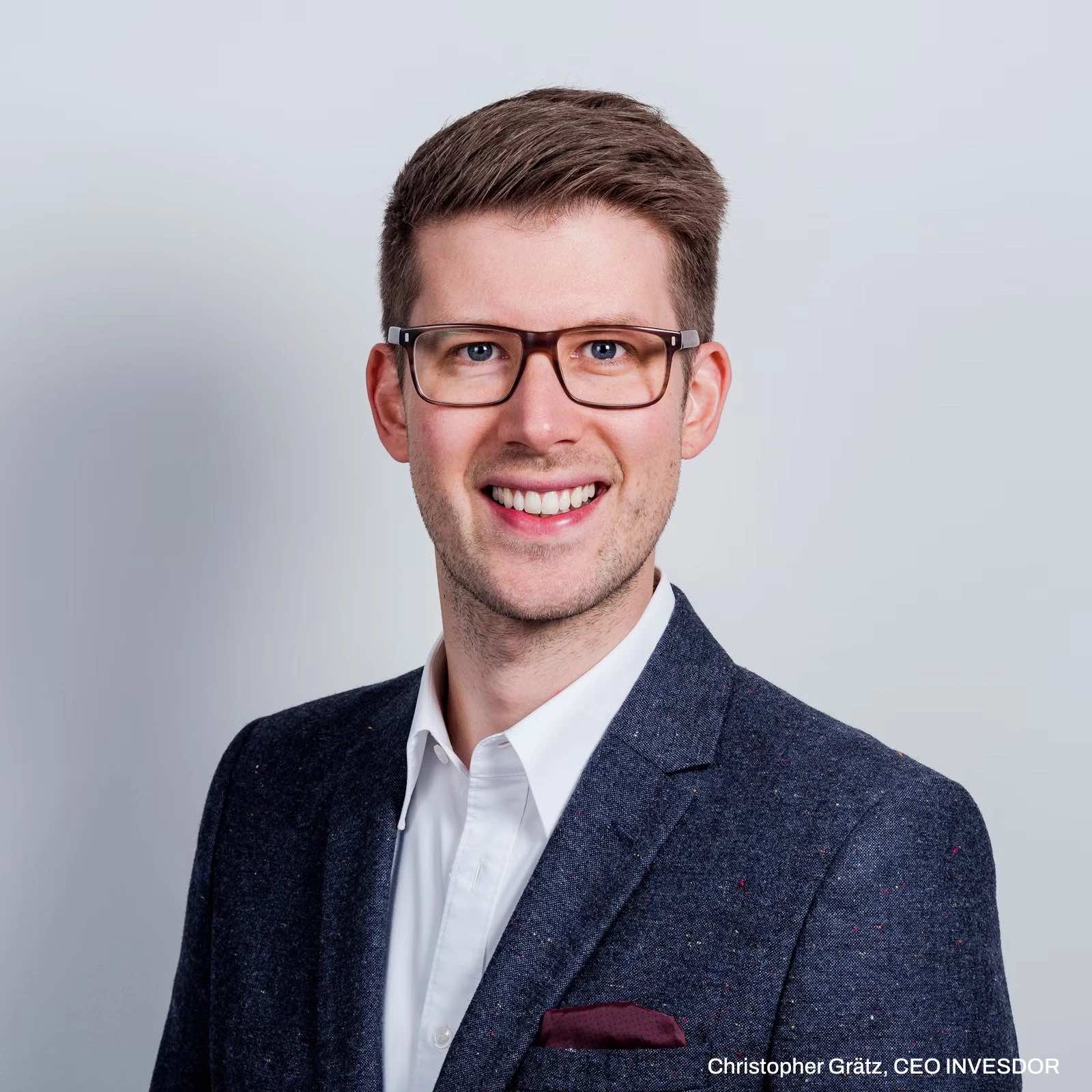 Ein Mann mit kurzen braunen Haaren und Brille lächelt, trägt einen dunklen Blazer, ein weißes Hemd und ein Einstecktuch und posiert vor einem hellgrauen Hintergrund. Der Text am unteren Rand lautet: Christopher Grätz, Vorstandsvorsitzender von Invesdor.