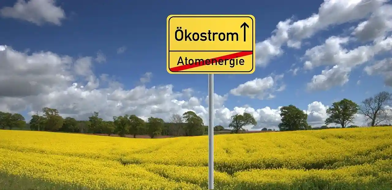 Auf einem gelben Straßenschild in einem gelben Blumenfeld steht Ökostrom mit einem Pfeil nach oben, und Atomenergie ist rot durchgestrichen, was den Wechsel von Kernenergie zu Ökostrom symbolisiert - genau wie die Wahl von Naturstrom Login für umweltfreundlichen Strom. Im Hintergrund sind Bäume und Wolken zu sehen.