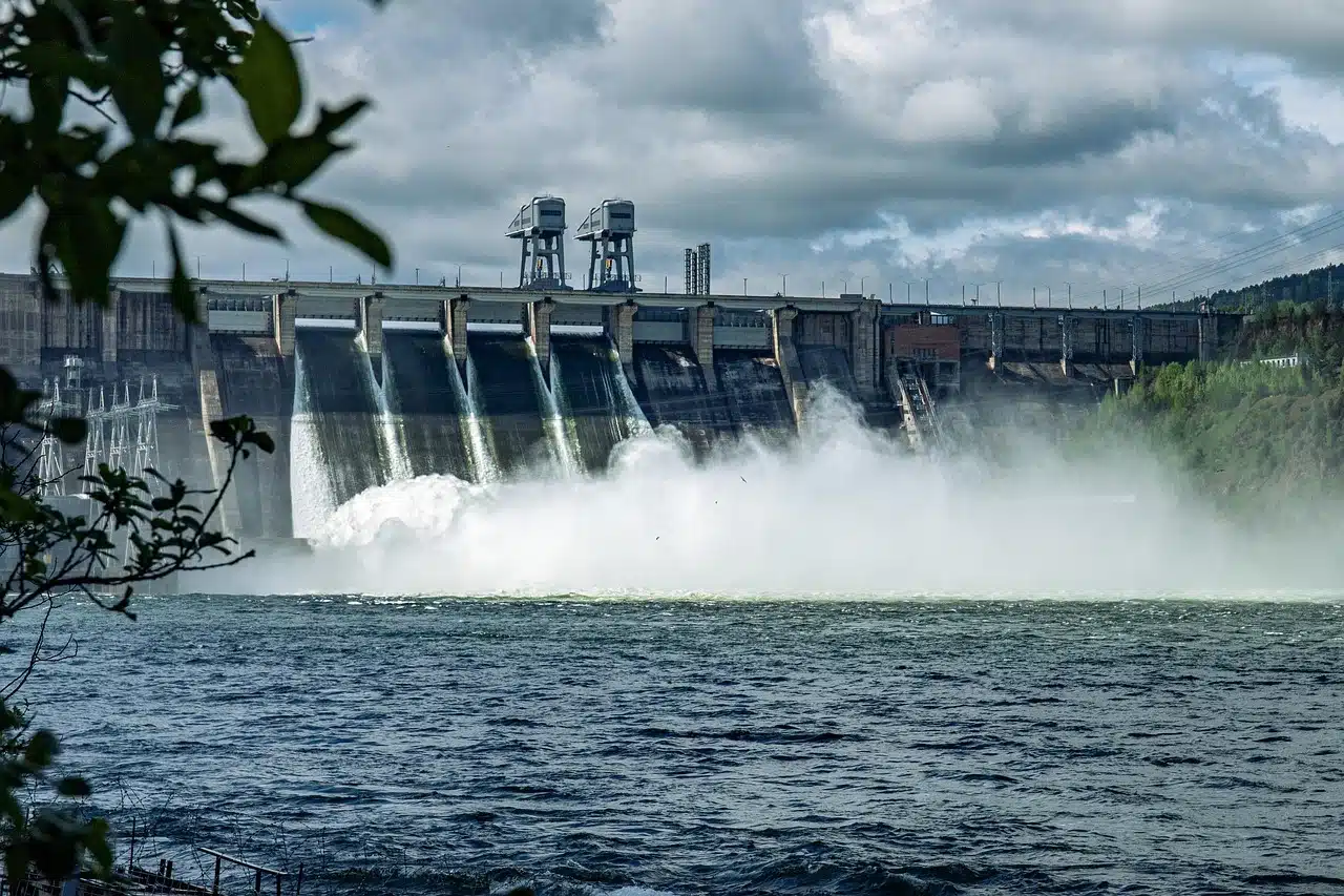Ein großer Wasserkraftdamm, der von Polarstern Strom angetrieben wird, lässt Wasser über mehrere Überlaufrinnen ab, wodurch Nebel und Turbulenzen im Fluss entstehen. Im Vordergrund ist grünes Laub unter einem bewölkten Himmel zu sehen.