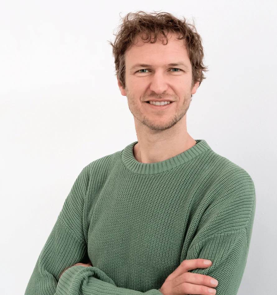 Ein lächelnder Mann mit gewelltem braunem Haar, der Klimakompetenz verkörpert, trägt einen hellgrünen Strickpullover und steht mit verschränkten Armen vor einem schlichten weißen Hintergrund.