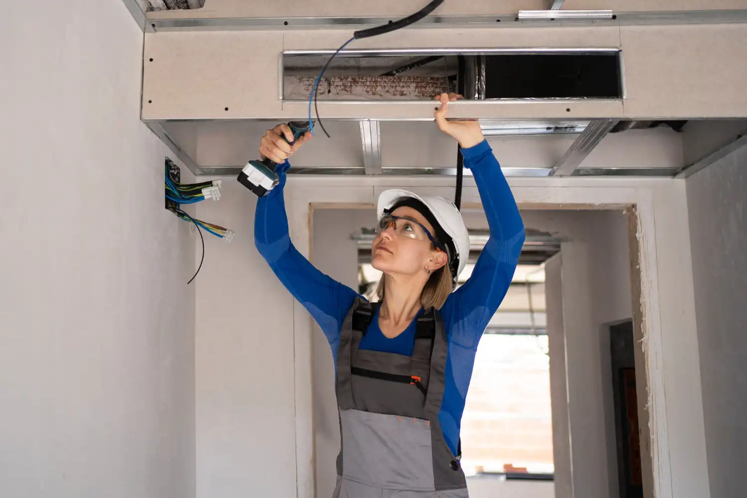 Eine Frau, die einen Schutzhelm und eine Schutzbrille trägt, arbeitet mit einer Bohrmaschine an elektrischen Leitungen in einer Decke, um energieeffiziente Lüftungssysteme in einem teilweise gebauten oder renovierten Raum vorzubereiten.