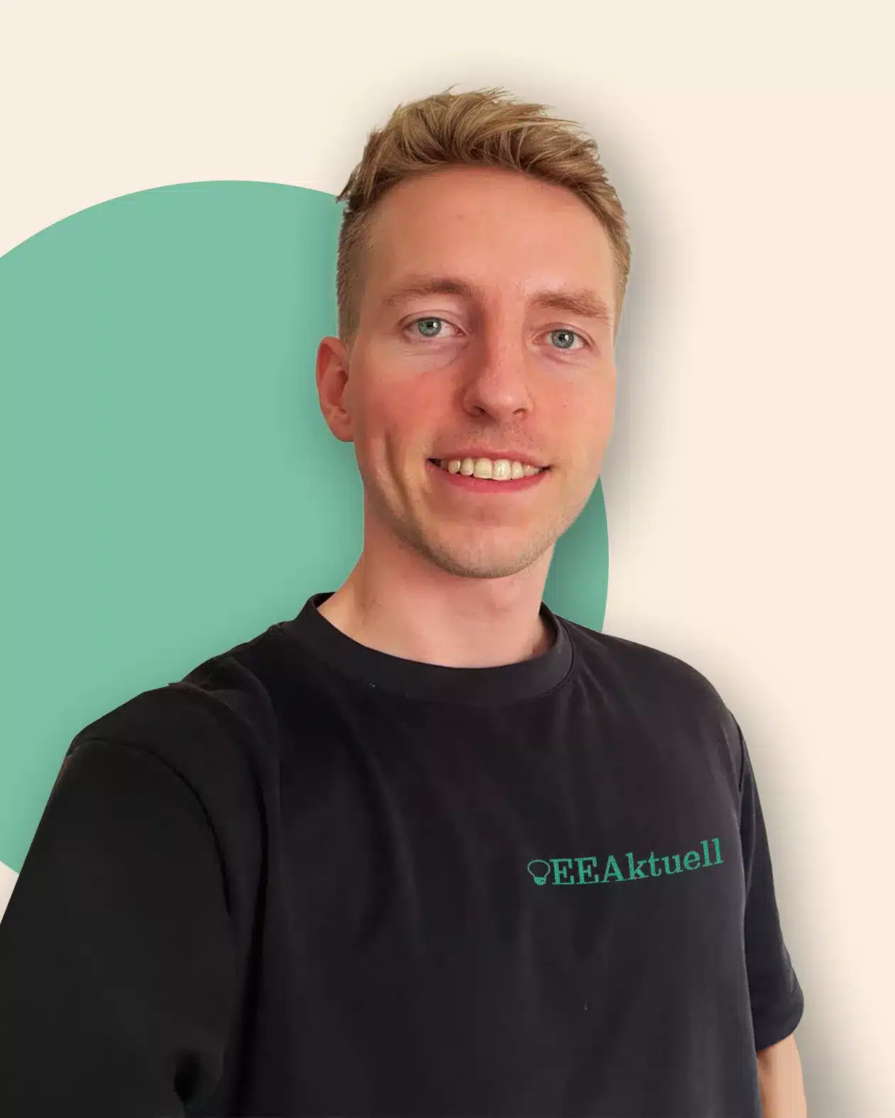Ein lächelnder junger Mann mit kurzen blonden Haaren trägt ein schwarzes "EEAktuell"-T-Shirt und steht vor einem cremefarbenen Hintergrund mit einem großen grünen Kreis, der das Thema Erneuerbare Energien hervorhebt.