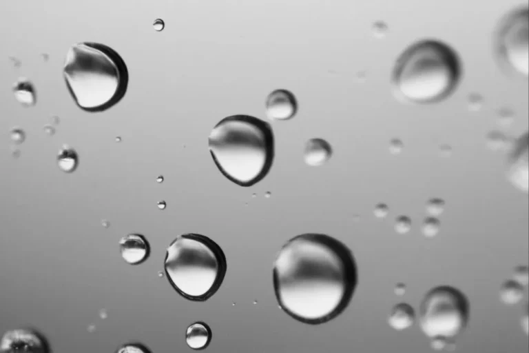 Nahaufnahme von Wassertröpfchen auf einer Glasoberfläche aus AISI 316L vor einem unscharfen grauen Hintergrund. Die Tröpfchen variieren in ihrer Größe und reflektieren das Licht, wodurch ein weiches, abstraktes Muster entsteht.