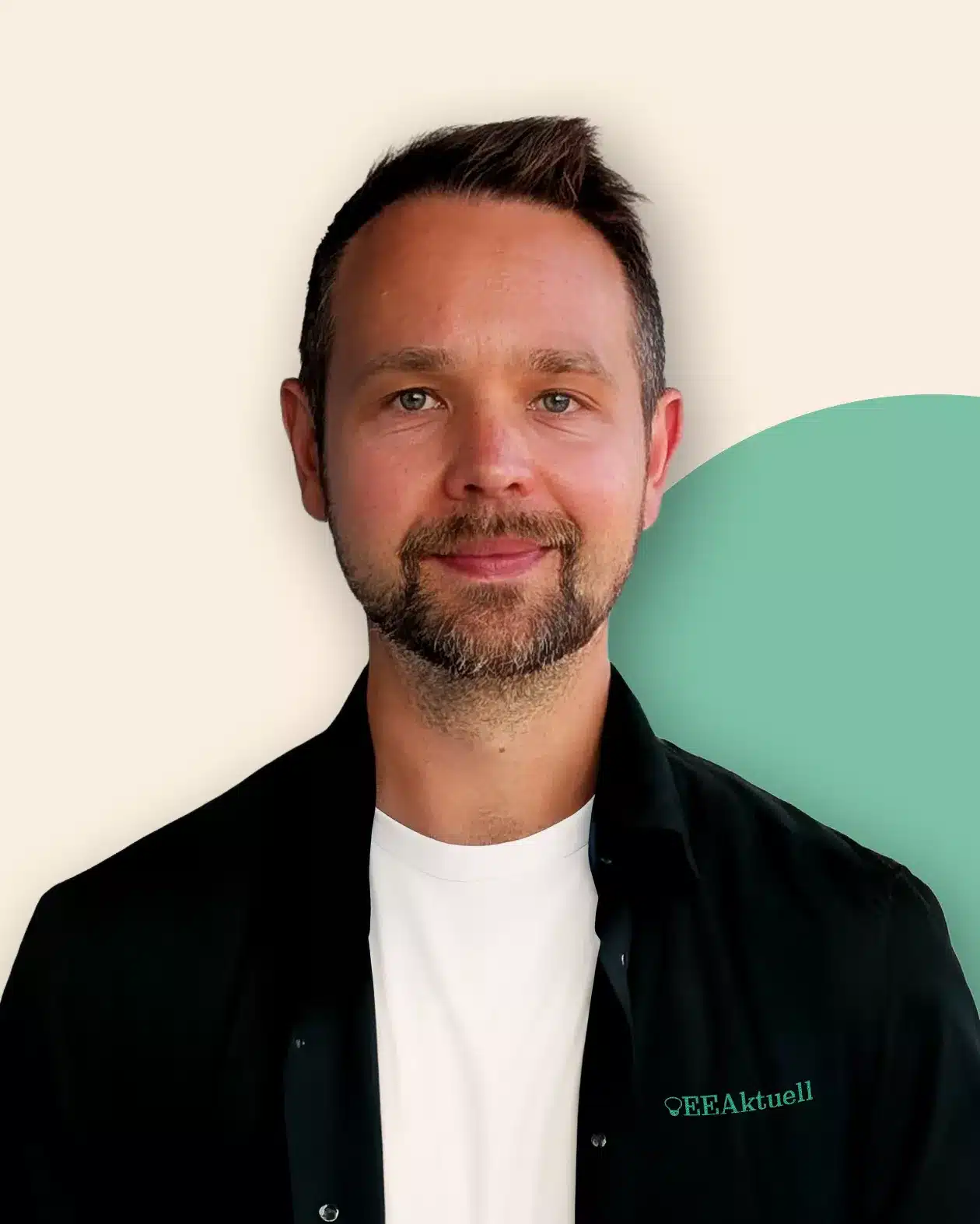 Ein Mann mit kurzen braunen Haaren und einem Bart lächelt leicht. Er trägt ein schwarzes Hemd über einem weißen T-Shirt mit der Aufschrift "OEEAktuell", was sein Interesse an Erneuerbaren Energien verdeutlicht. Der Hintergrund ist beige mit einem grünen, kreisförmigen Akzent.