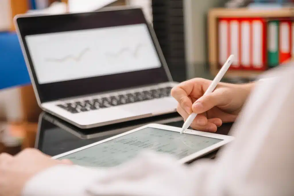 Eine Person benutzt einen Stift, um auf einem Tablet zu schreiben oder zu zeichnen, während im Hintergrund ein Laptop mit ETF für Anfänger-Diagrammen angezeigt wird. Die Szene suggeriert Datenanalyse oder digitale Arbeit in einer modernen Büroumgebung.